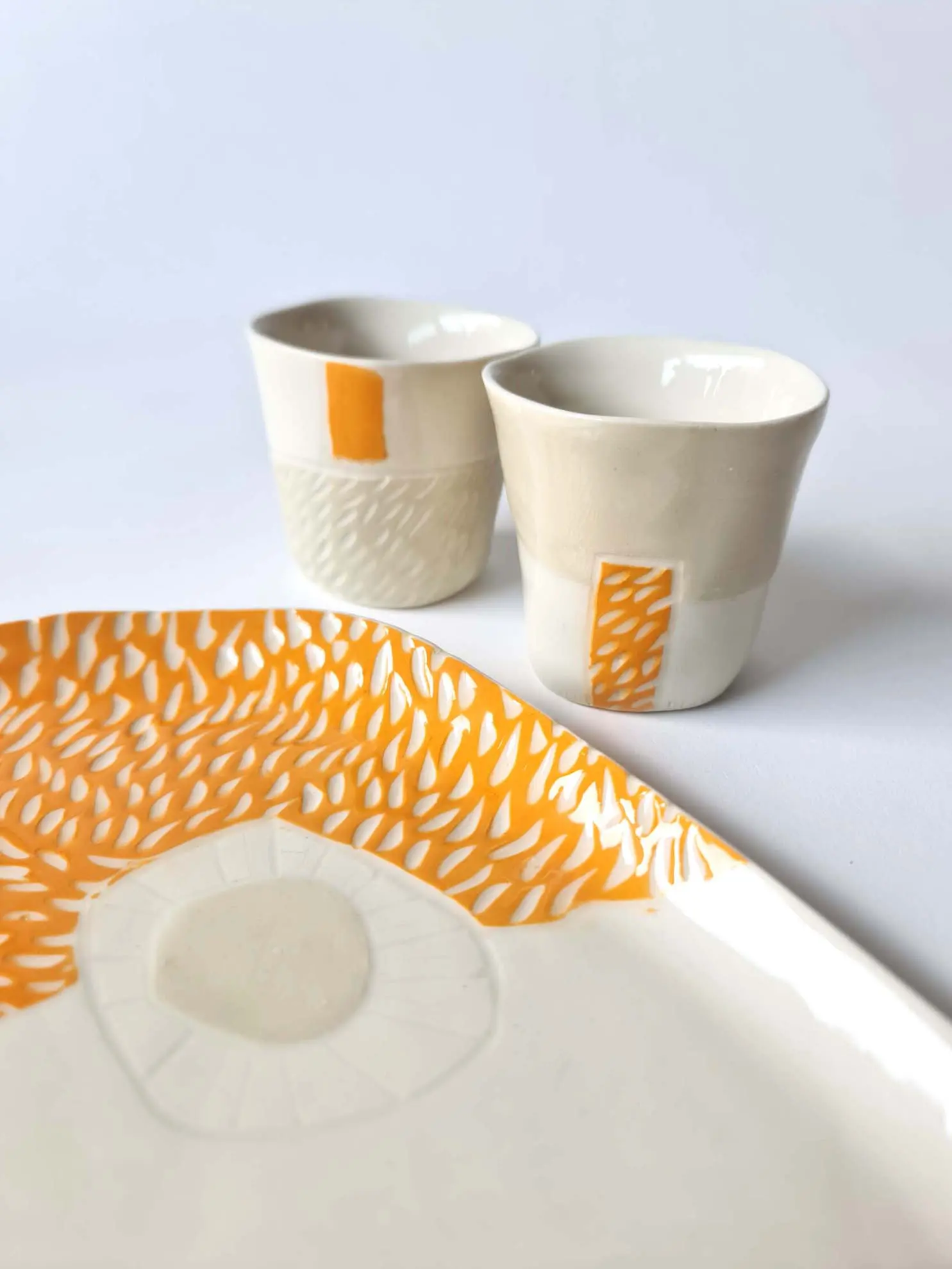 Handmade porcelain tray and two cups with orange sgraffito detailing on a white background.