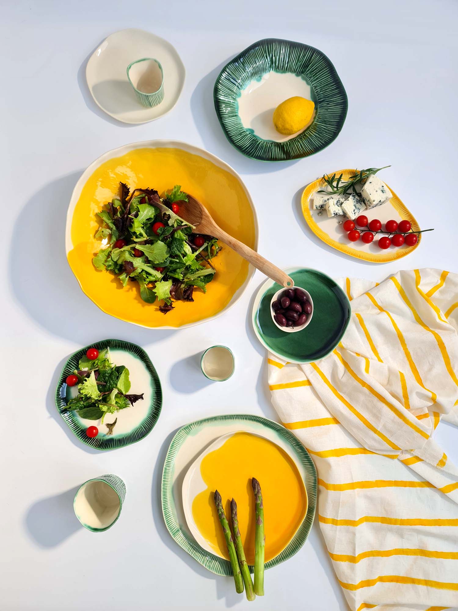 Colorful porcelain tableware with salad, asparagus, cheese, and lemons, styled on a white background with a yellow-striped cloth.