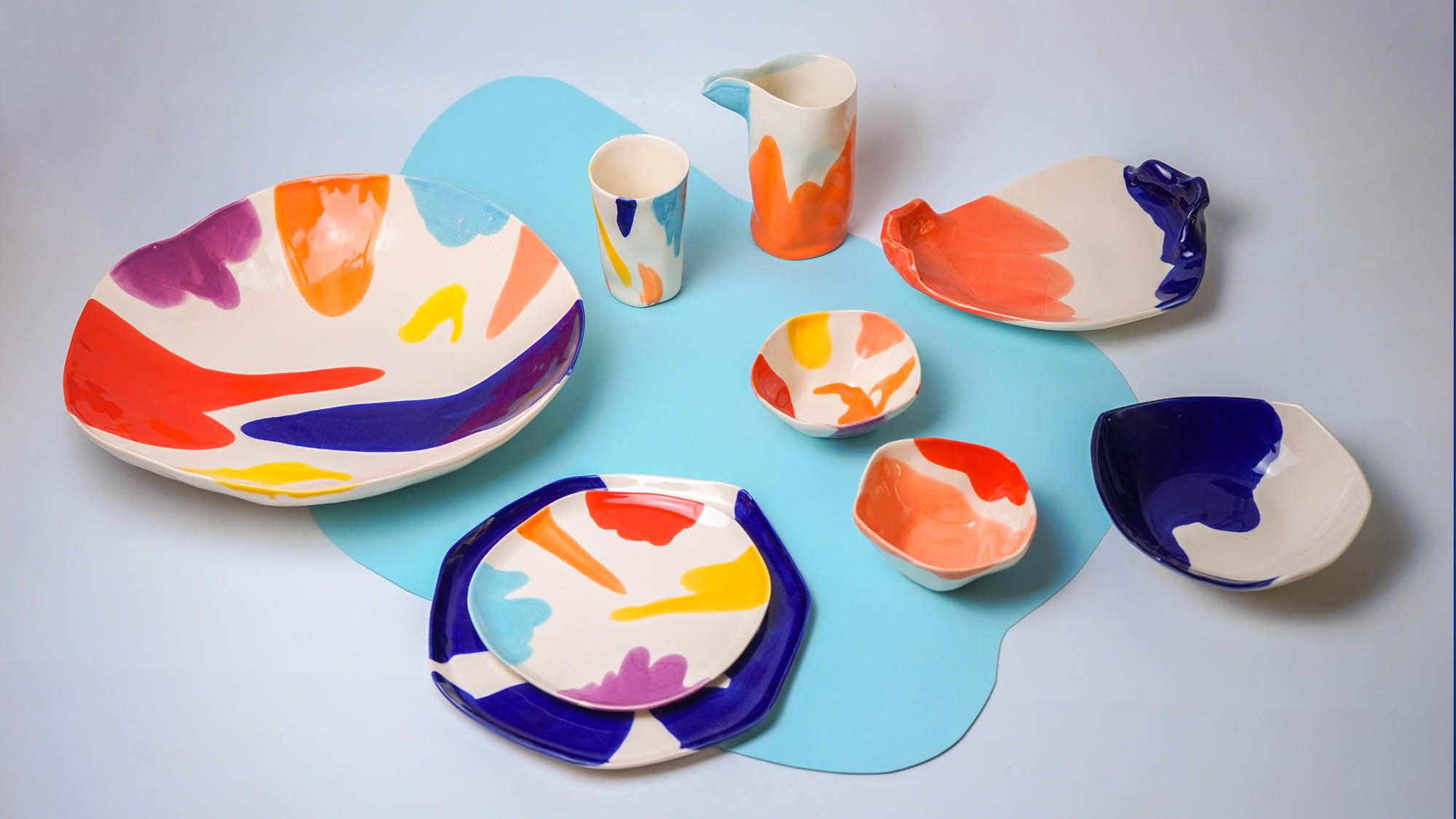 Colorful porcelain plates, bowls, and cups with multicolored brushstroke designs on a light blue background.