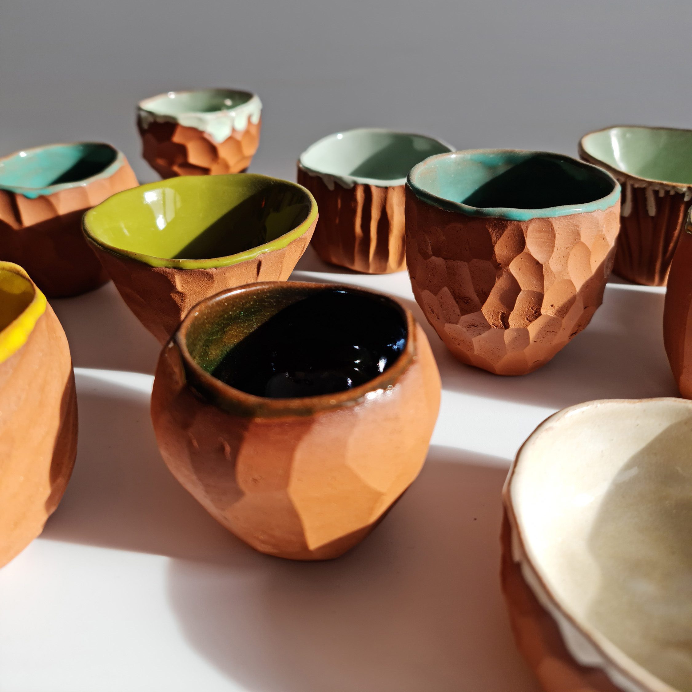 A close-up of a small cup from the Painted Ruins collection. The vessel is handcrafted from terracotta, featuring a chiseled, faceted exterior left unglazed to highlight the raw texture of the clay. The rim is softly irregular, with a glossy green glaze cascading slightly over the edge and covering the interior.