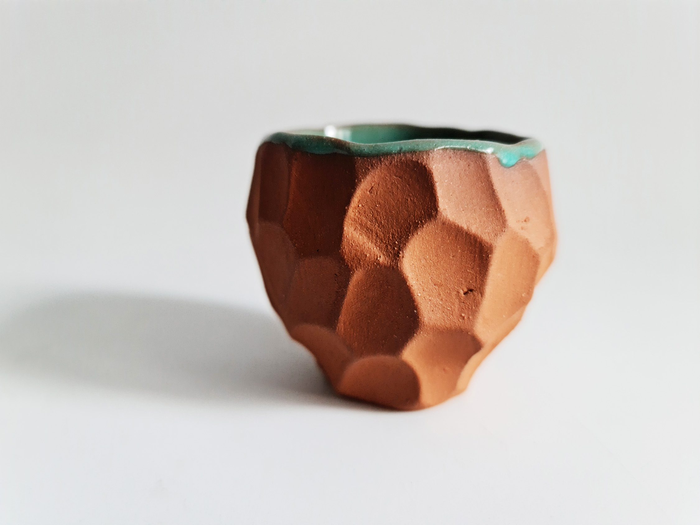 Faceted terracotta cup with a green glazed interior on a white background.