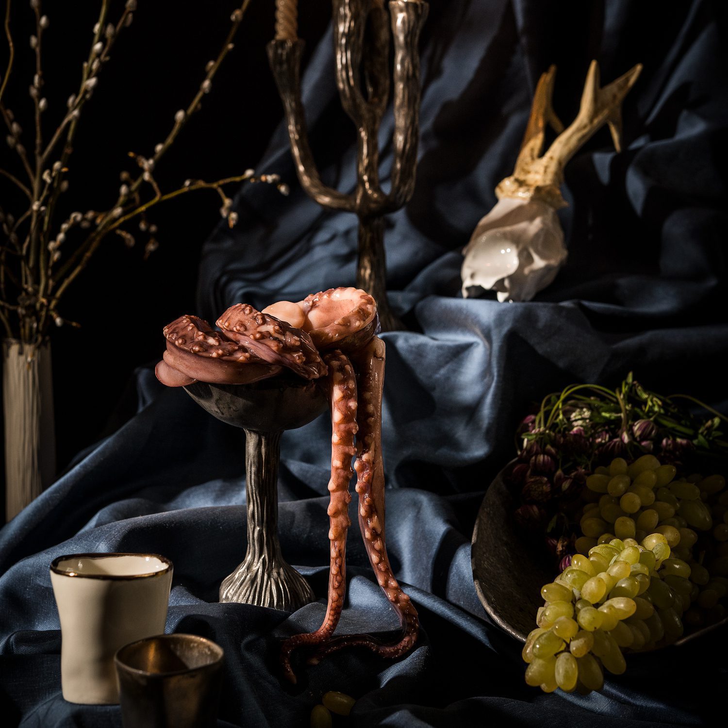 Dramatic still life with octopus tentacles on a sculptural ceramic pedestal, grapes, candles, and dark drapery.