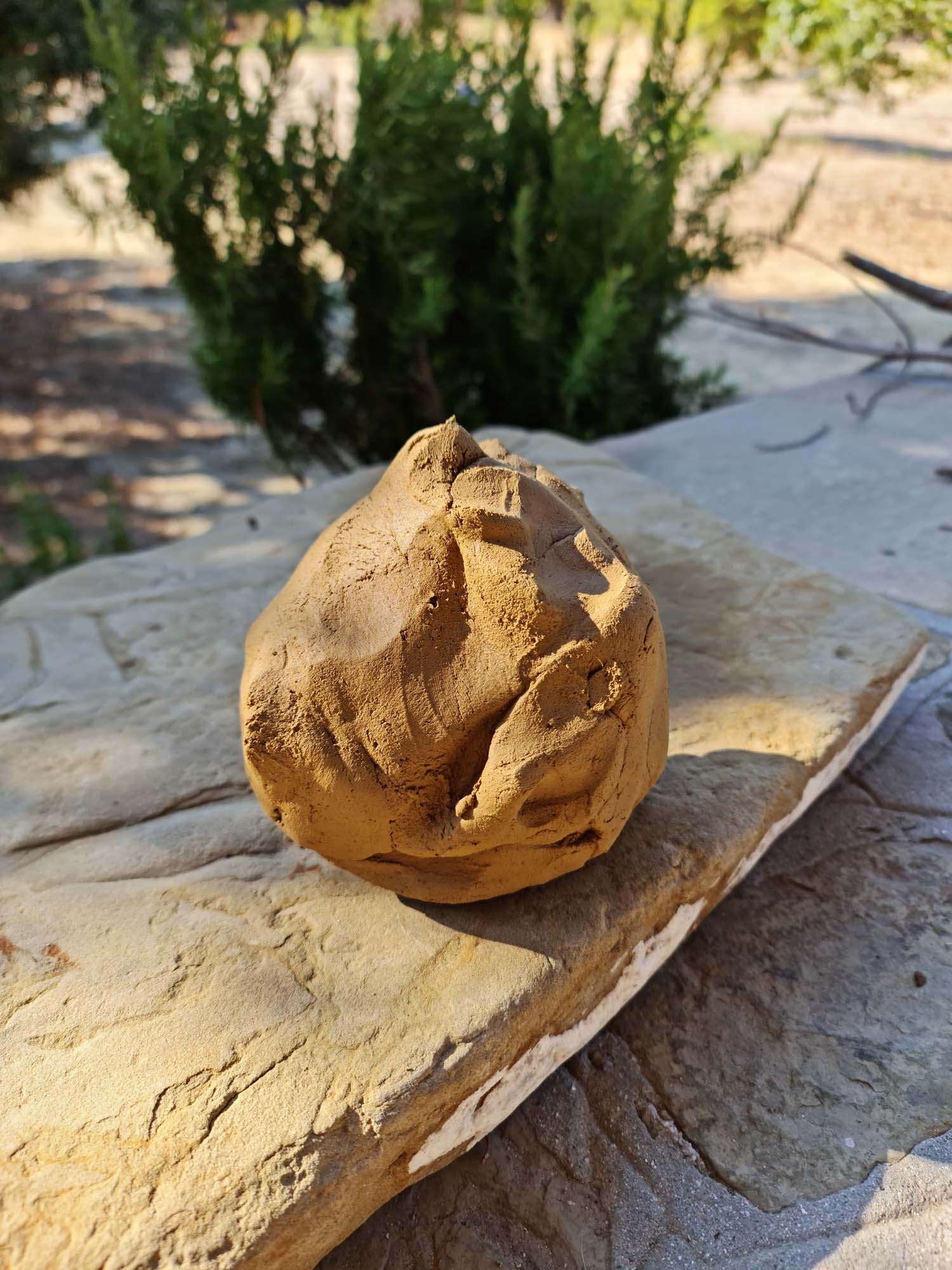 A lump of wild clay resting on a flat stone surface, with greenery in the background.